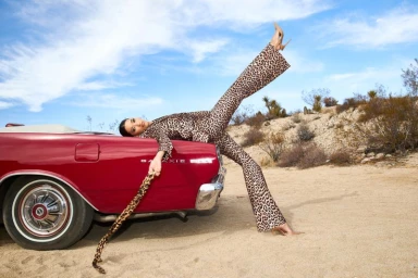 Model lying on the back of a vintage red convertible in a desert setting wearing a leopard print pantsuit