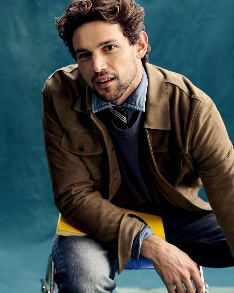Portrait of a dark-haired man in a brown jacket and jeans sitting on a blue stool with a yellow book against a teal background.