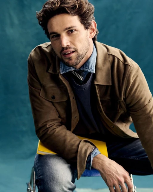 Portrait of a dark-haired man in a brown jacket and jeans sitting on a blue stool with a yellow book against a teal background.