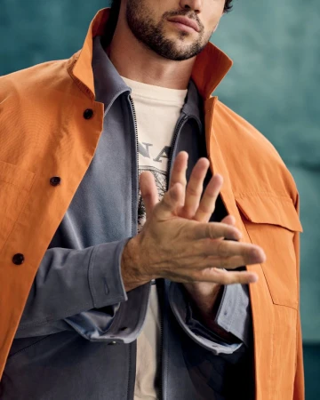 Man rubbing hands together wearing an orange jacket, grey shirt, and white t-shirt against a textured teal background