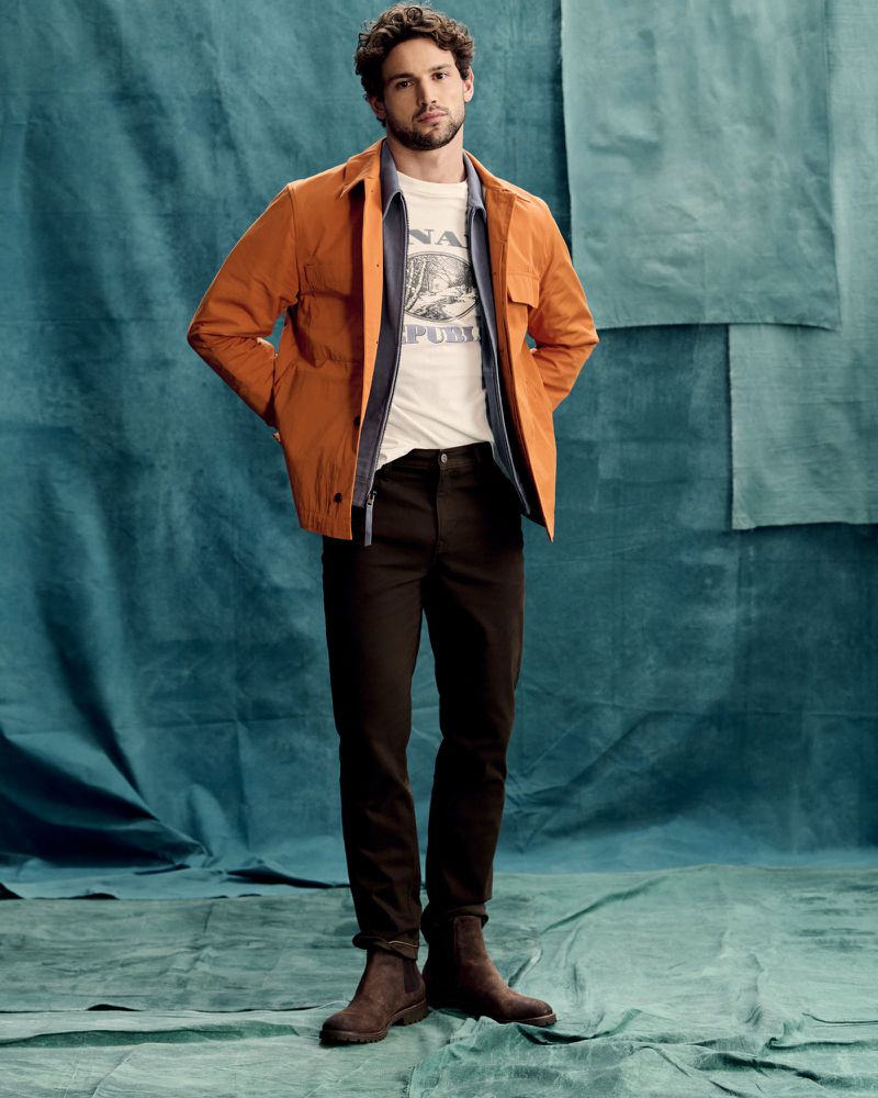 Full-length shot of adult man posing in an orange work jacket, t-shirt, dark pants, and boots against a blue textured background.