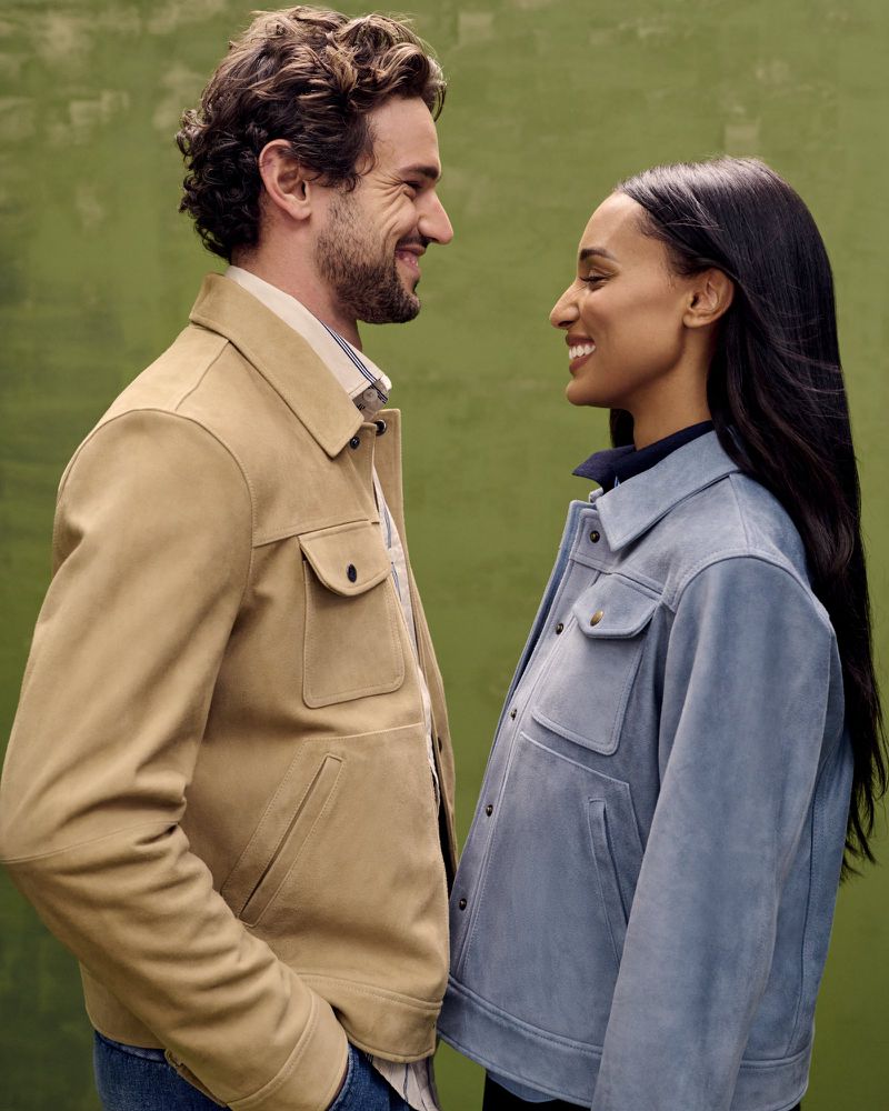 A smiling couple wearing matching jackets, a man in a tan jacket and a woman in a light blue jacket, standing against a green backdrop.