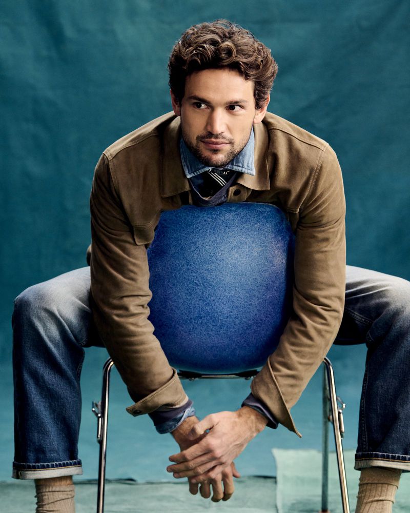 Portrait of a man in a brown jacket and jeans sitting backward on a blue chair in a photo studio with a blue background.