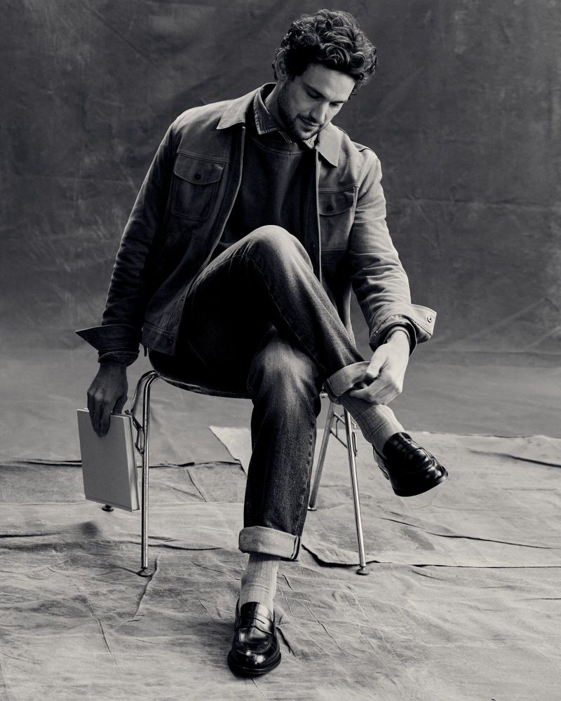 Monochrome portrait of an adult male sitting on a chair, rolling up his jeans cuff. He is holding a book. Studio setting.