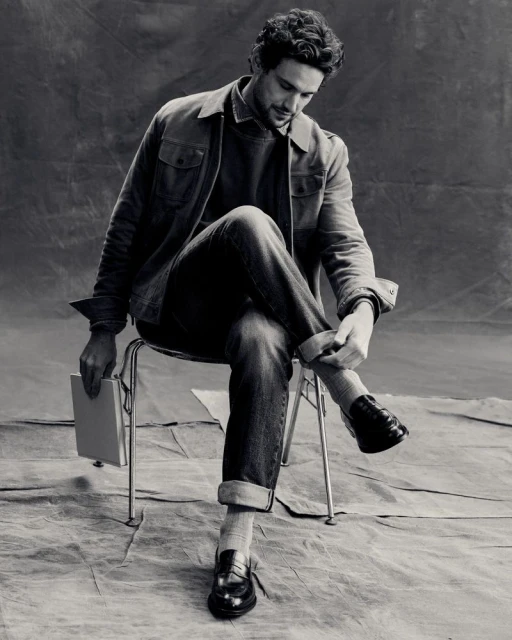 Monochrome portrait of an adult male sitting on a chair, rolling up his jeans cuff. He is holding a book. Studio setting. 