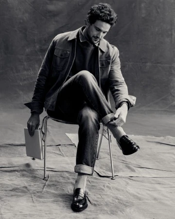 Monochrome portrait of an adult male sitting on a chair, rolling up his jeans cuff. He is holding a book. Studio setting. 