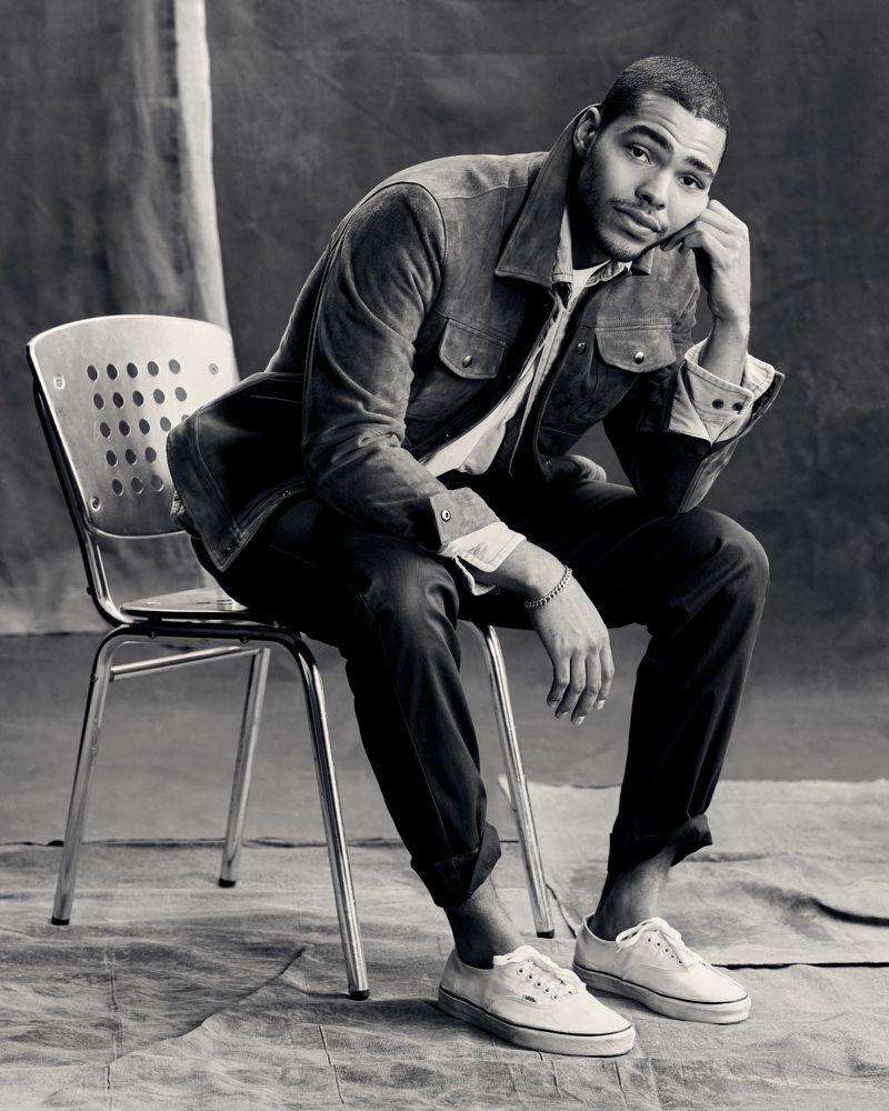 Man in a jacket sitting on a metal chair in front of a textured background. He wears jeans and white sneakers in a studio setting.