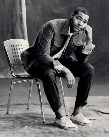 Man in a jacket sitting on a metal chair in front of a textured background. He wears jeans and white sneakers in a studio setting.