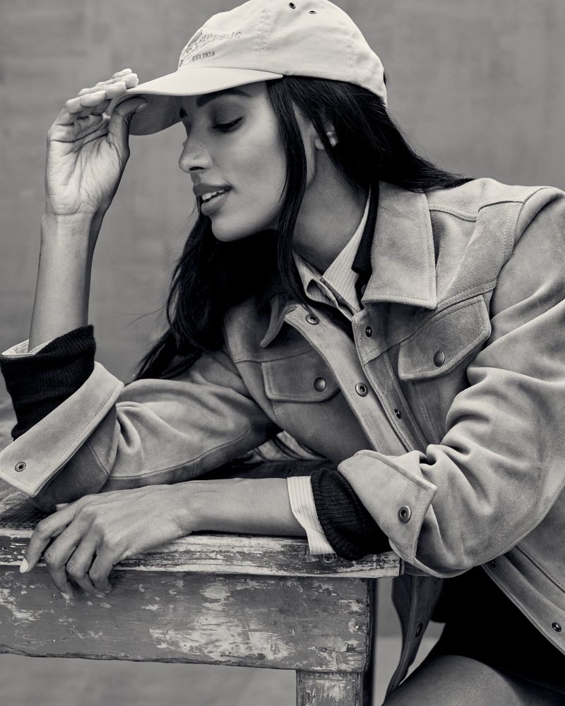Black and white portrait of a woman wearing a suede jacket and baseball cap, resting on a rustic wooden table.