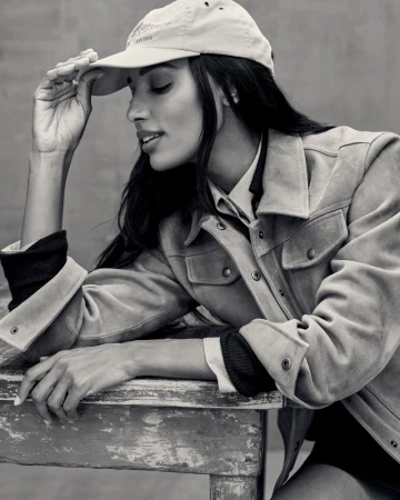Black and white portrait of a woman wearing a suede jacket and baseball cap, resting on a rustic wooden table.