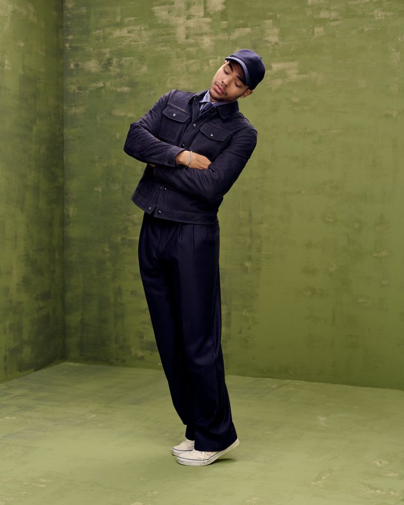 Male fashion model posing in an indoor studio with a textured green wall, wearing a navy jacket, trousers, cap, and white shoes.