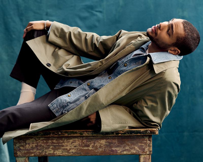 A man with a beard sits on a weathered wooden bench, wearing a khaki trench coat, denim jacket, and black pants against a teal backdrop.