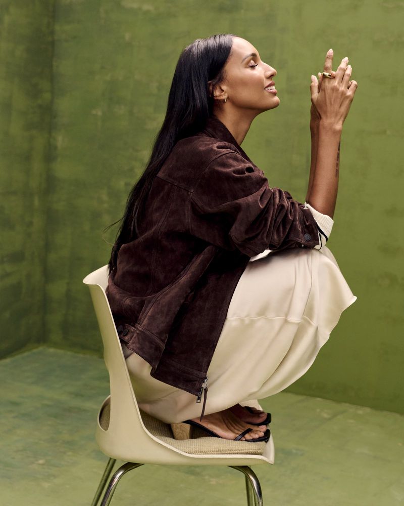 A woman with long dark hair sits on a chair indoors, wearing a brown jacket, cream-colored pants, and sandals against an olive-green wall.