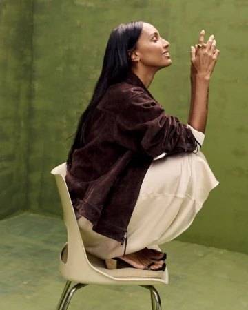 A woman with long dark hair sits on a chair indoors, wearing a brown jacket, cream-colored pants, and sandals against an olive-green wall.