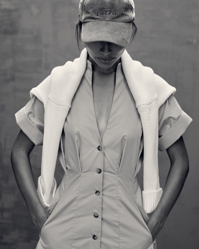 Woman in a dress and baseball cap with sweater draped over her shoulders, posing indoors with a gray background.