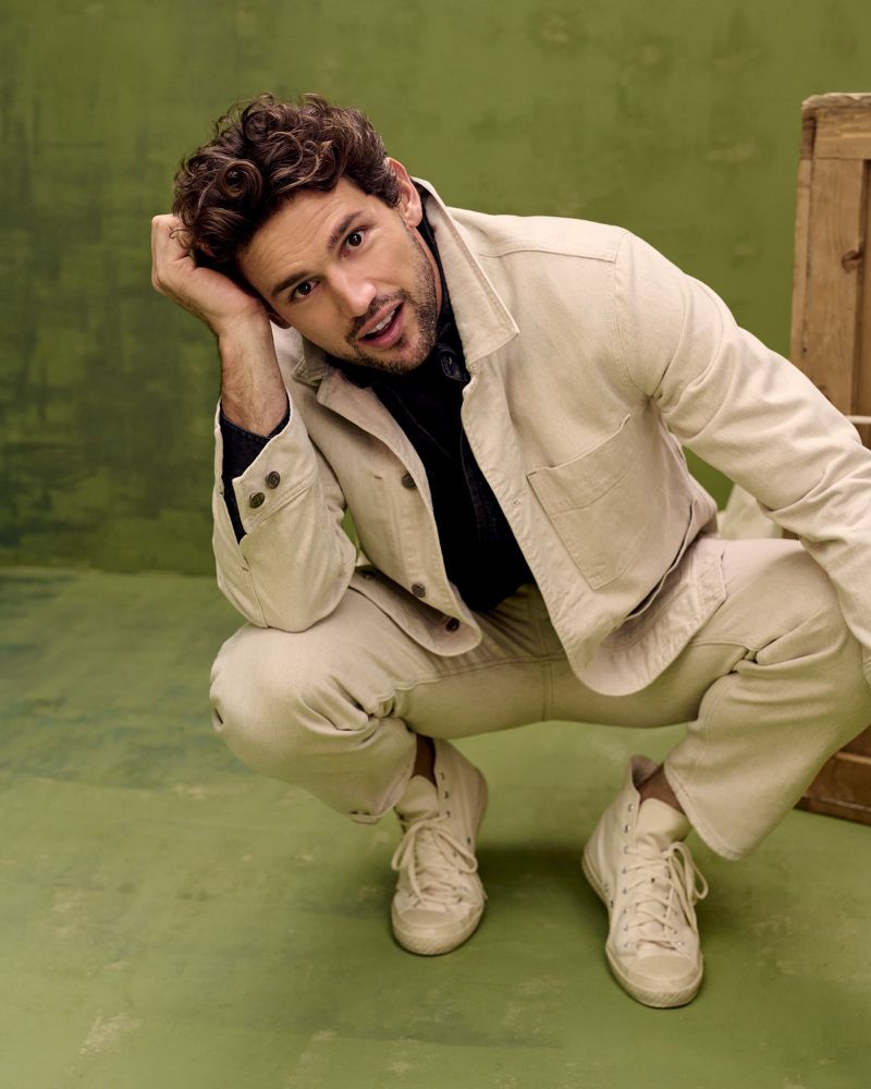 Handsome man with curly hair in light-tan jacket and pants crouches in a studio setting against a mottled green backdrop.