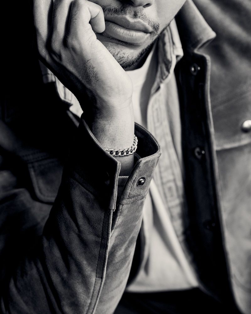 Portrait of a light-skinned adult male in a button-down shirt and suede jacket resting his chin on his hand displaying a chain bracelet.