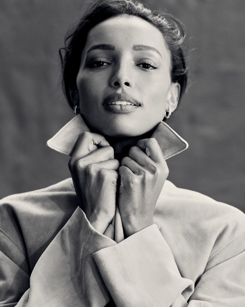 Black and white portrait of an adult female with her hands near her face, wearing a jacket with a wide collar, set against a gray background.