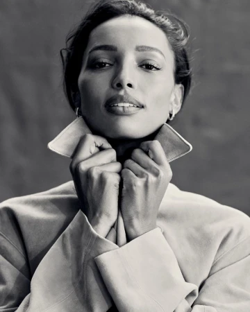 Black and white portrait of an adult female with her hands near her face, wearing a jacket with a wide collar, set against a gray background.