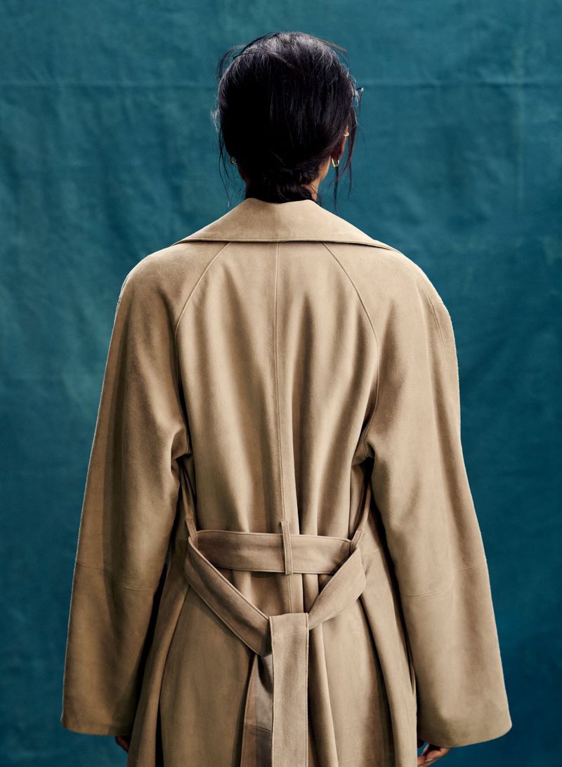 Rear view of a woman wearing a camel trench coat with a belted waist against a dark teal textured backdrop