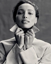 Close-up portrait of model Jasmine Tookes wearing a collared coat, adjusting the collar against a textured dark background.