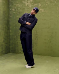 Full-length portrait of a male model wearing a dark navy jacket, trousers, and baseball cap against a green textured studio background.