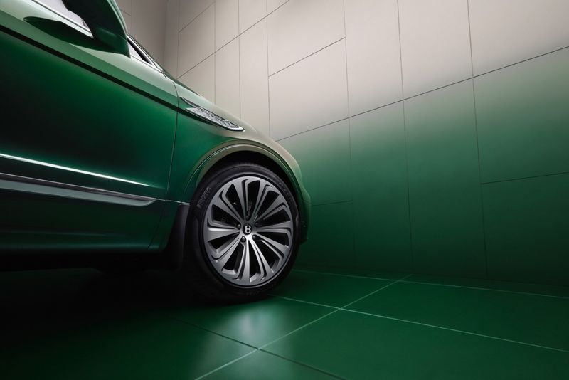 Close-up of a green Bentley SUV focusing on the wheel and front panel with a gradient white-to-green wall in the background.