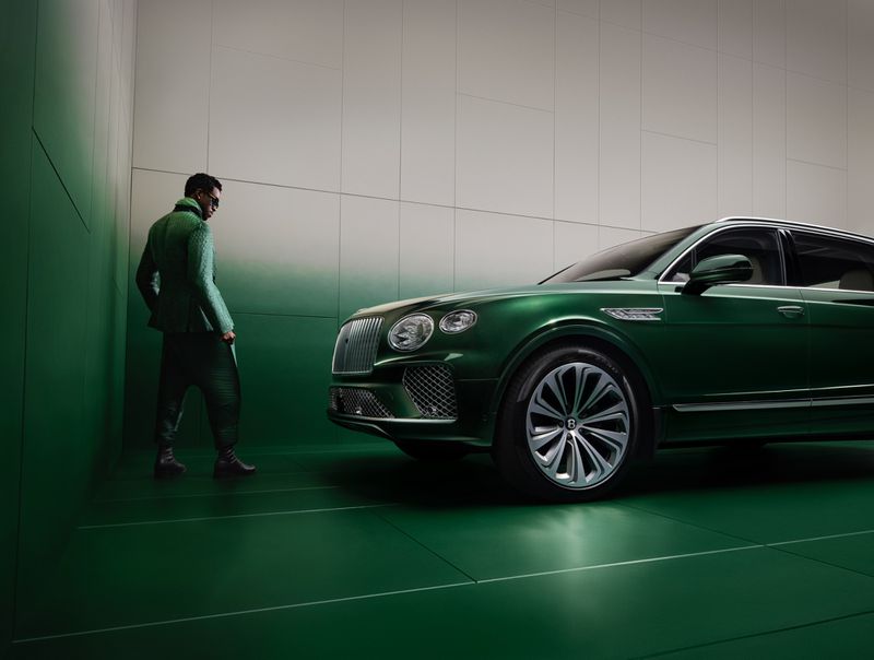 A green Bentley SUV is parked indoors with a man in a green suit standing nearby, all set against a green and white wall.