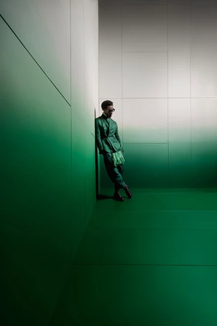 A man in a green suit stands in a corner of a room with a green floor and green and white walls, under studio lighting.