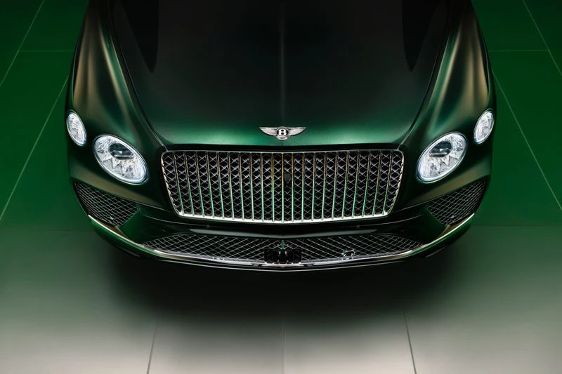 The front of a glossy dark green Bentley car is showcased with its iconic grill, headlights, and emblem against a graduated green backdrop.