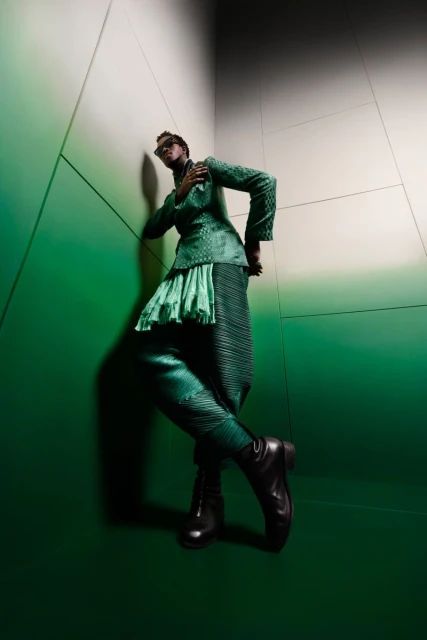 A young adult fashion model in emerald green stands in a corner with structured clothing, accessorized with black leather boots and sunglasses.