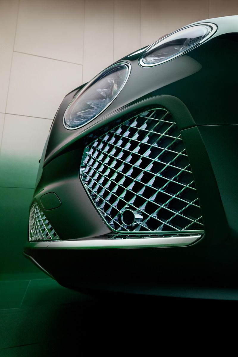 Close-up of the front of a dark green Bentley showcasing its headlight, grill, and front bumper against a light grey background.
