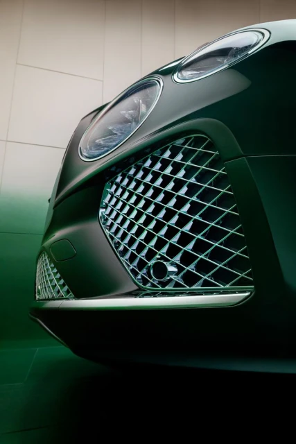 Close-up of the front of a dark green Bentley showcasing its headlight, grill, and front bumper against a light grey background.