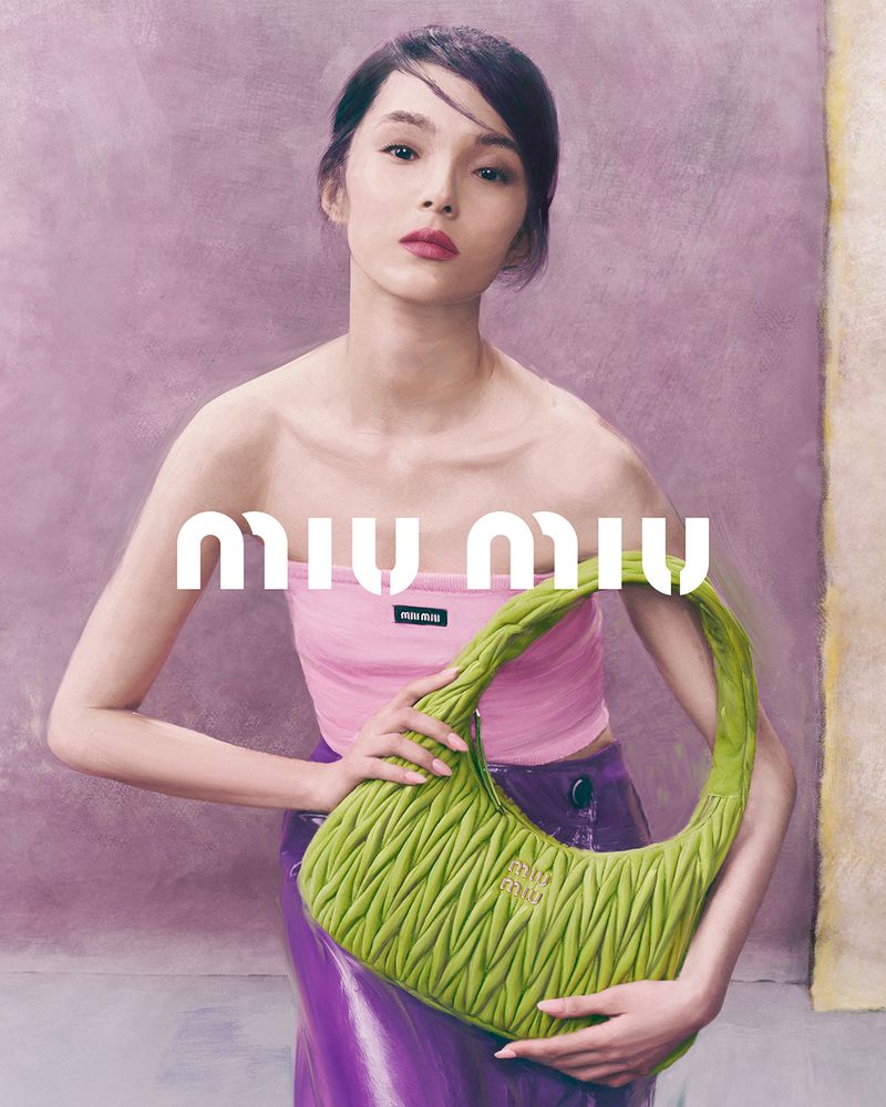 Fashion shot of an Asian woman posing in a pink top and purple skirt, holding a green Miu Miu handbag against a textured backdrop.