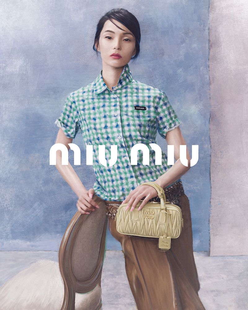 Fashion shot of a woman in a blue and green checkered shirt holding a beige handbag in front of a blue painted backdrop.