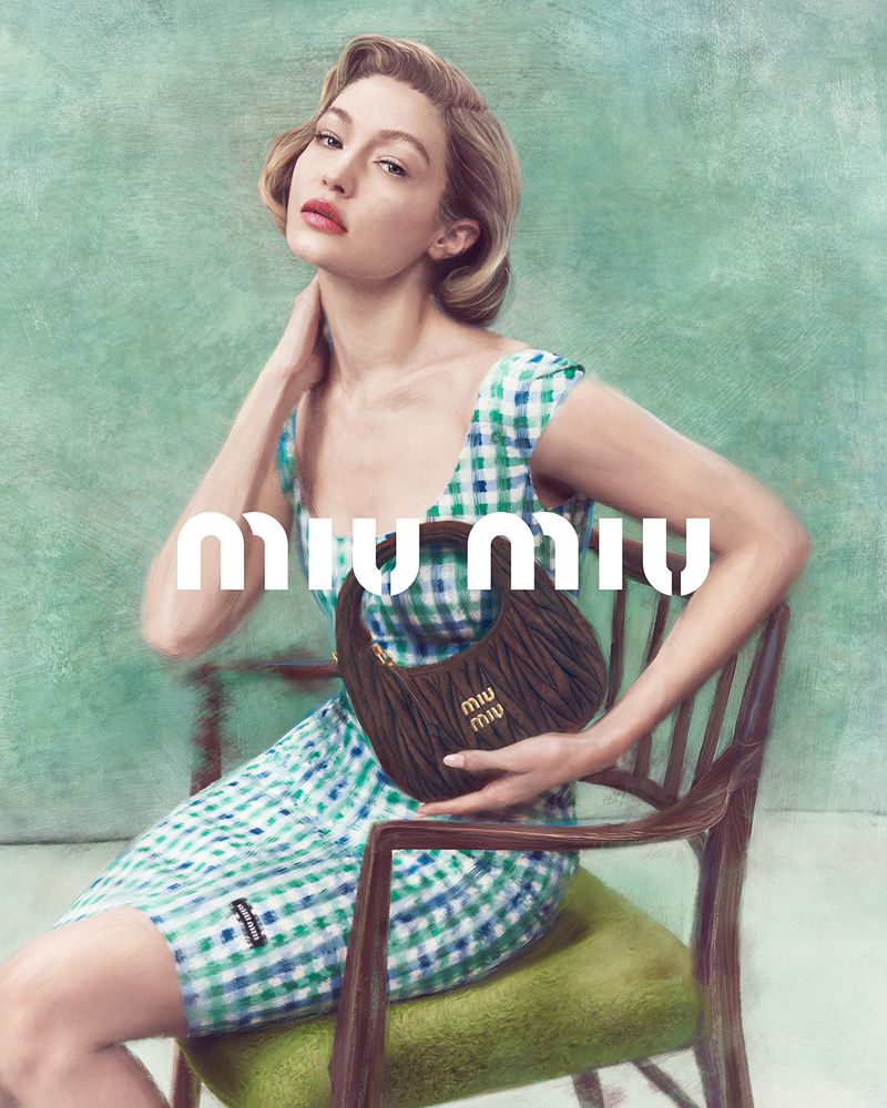 Gigi Hadid models a Miu Miu purse, seated on a chair in front of a mottled green backdrop while wearing a blue and white dress.