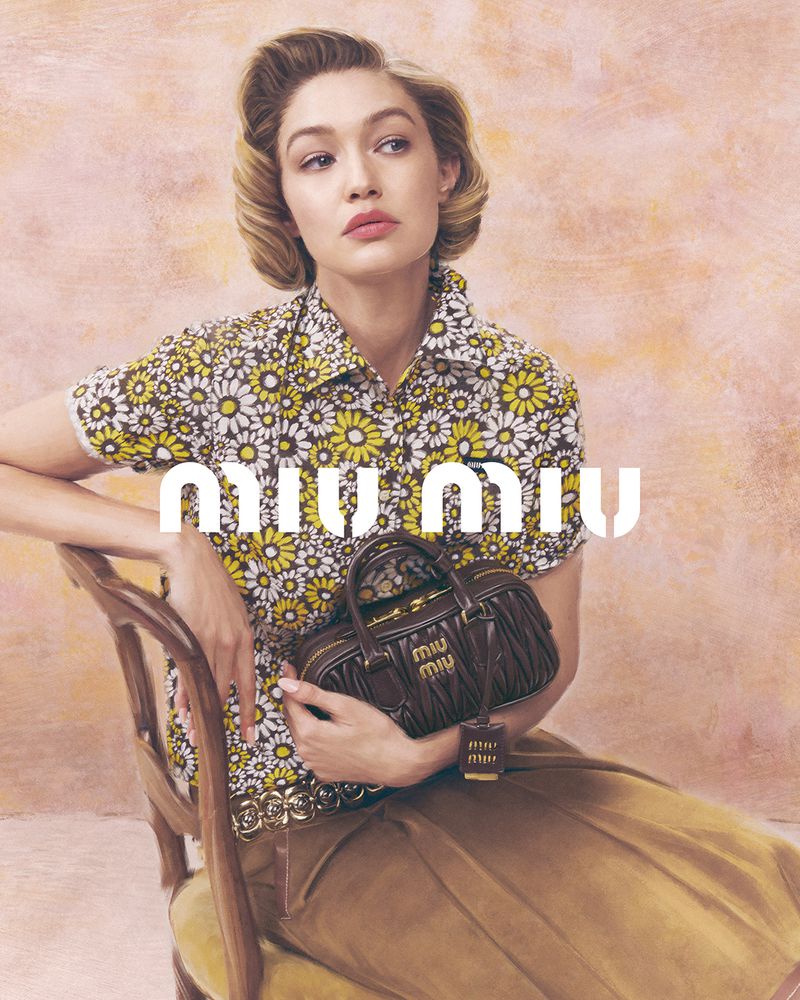Gigi Hadid sits with a Miu Miu purse in a fashion campaign shot. She wears a flower-print top and tan skirt against a textured backdrop.