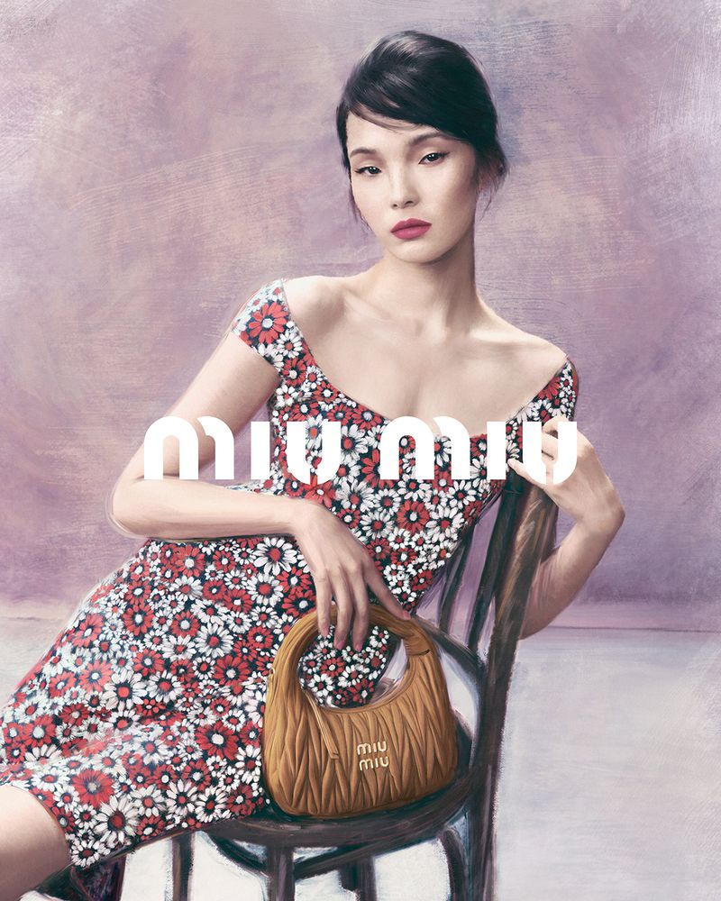 Model Xiao Wen Ju seated on a wooden chair wearing a floral dress holding a brown leather handbag for Miu Miu
