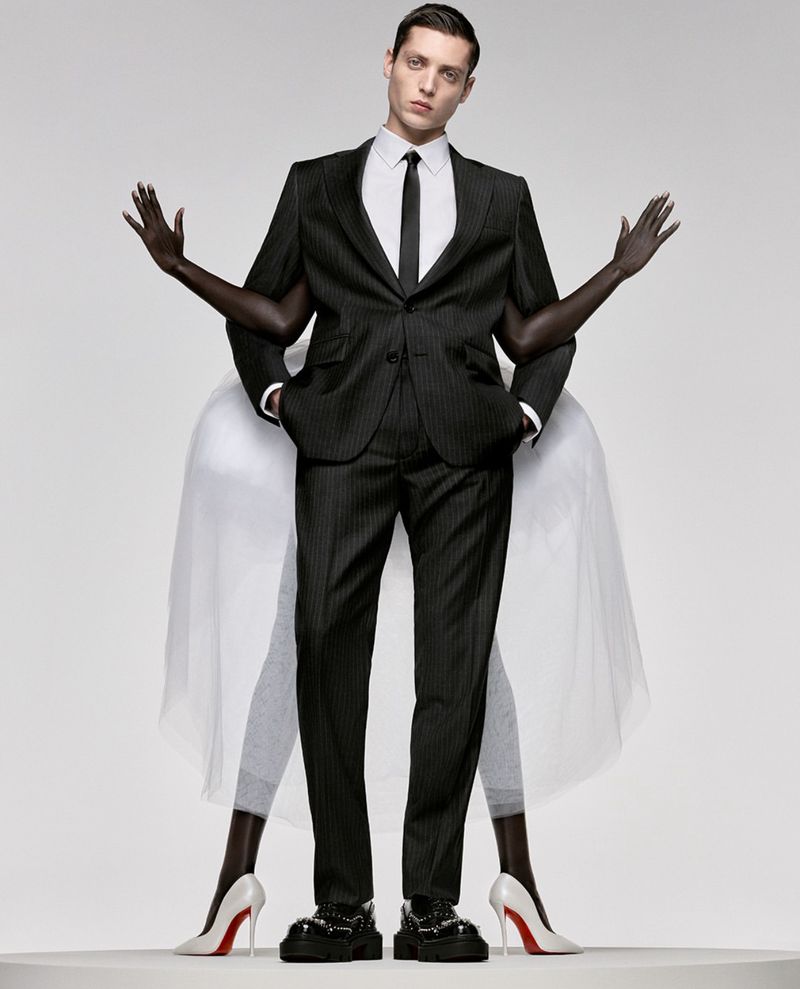 Man in a pinstripe suit stands centered in front of woman's arms and legs in a studio setting on a white background.