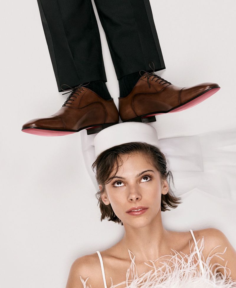 Woman looks up at a man's shoes; she wears a feathered white top and cap while lying on a white surface, creating a unique portrait.