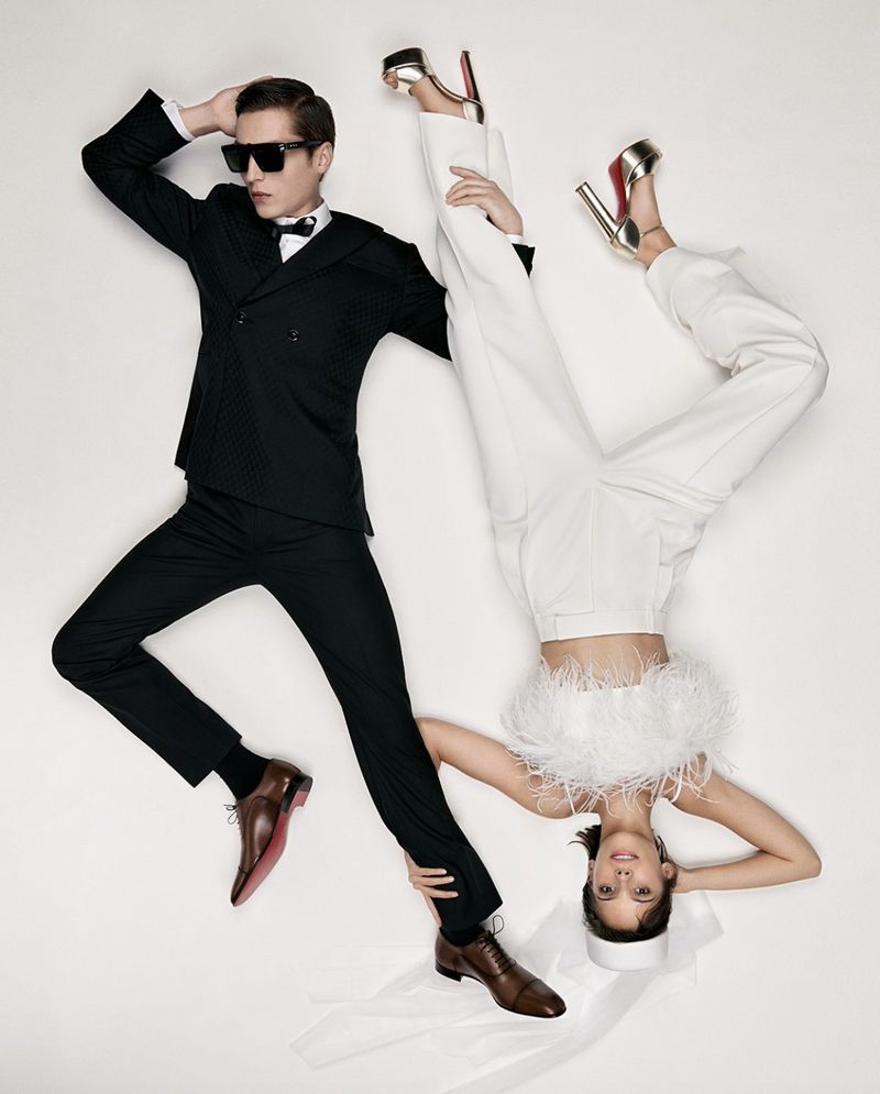 Fashion shot of a man in a black tuxedo and a woman in a white outfit with feather trim, posing together on a white background.