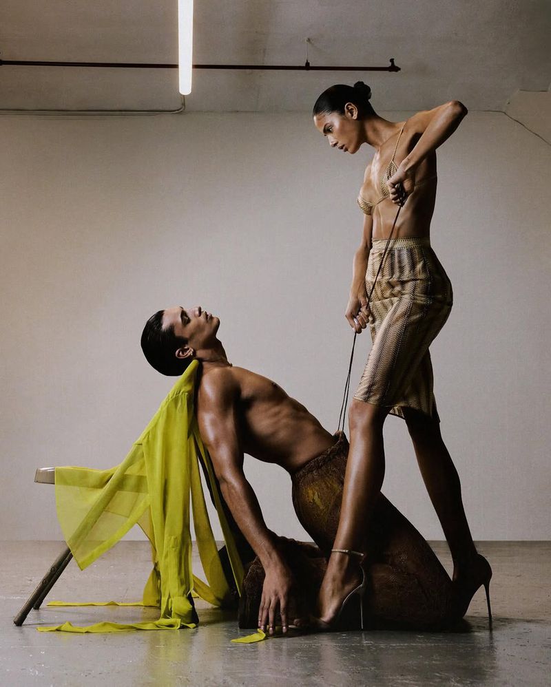Two slender adults in a studio, one in heels standing over the other who is seated on a chair draped in a semi-transparent yellow fabric.