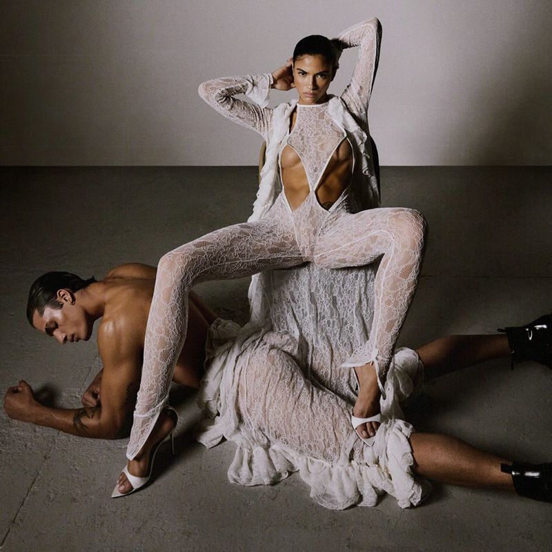 A tanned woman in a cutout lace jumpsuit poses, seated on a man wearing black boots. The photo is set against a neutral studio backdrop.