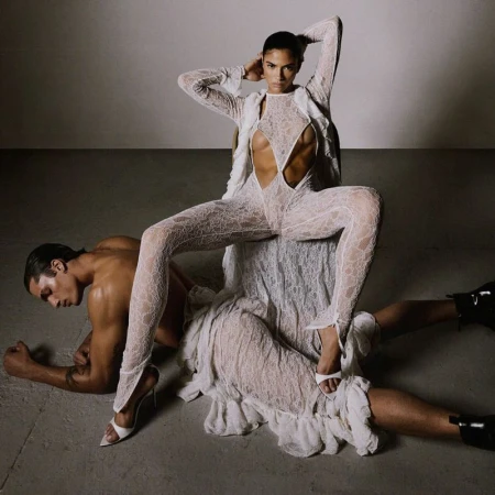 A tanned woman in a cutout lace jumpsuit poses, seated on a man wearing black boots. The photo is set against a neutral studio backdrop.