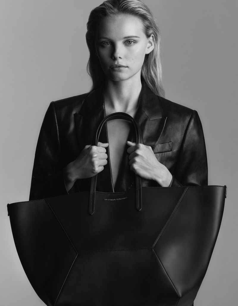 Black and white studio shot of a blonde woman in a blazer holding a large geometric handbag, showcasing fashion and style.