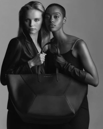 Two models pose in a black and white studio shot, showcasing a large black leather tote bag, both exuding a serious expression.