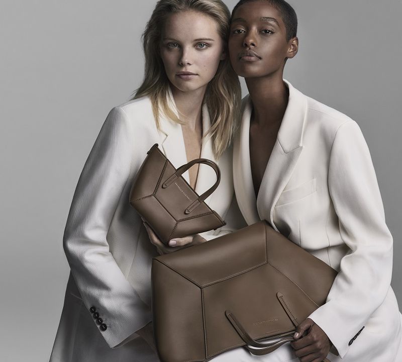 Two female models wearing white blazers holding brown leather handbags against a grey backdrop
