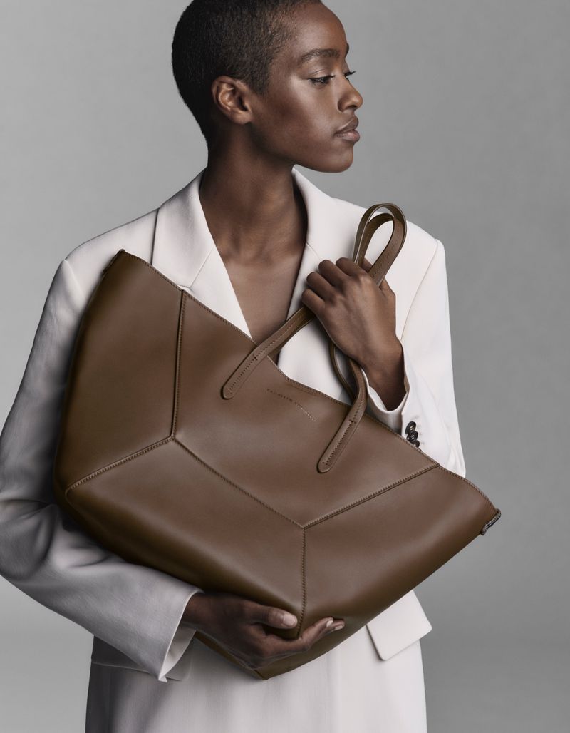 Black model holding a large brown leather tote bag wearing an off-white blazer against a neutral grey studio background.