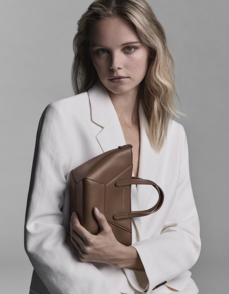 Fashion portrait of a blonde adult woman holding a tan angular handbag, wearing a white suit jacket against a gray background.