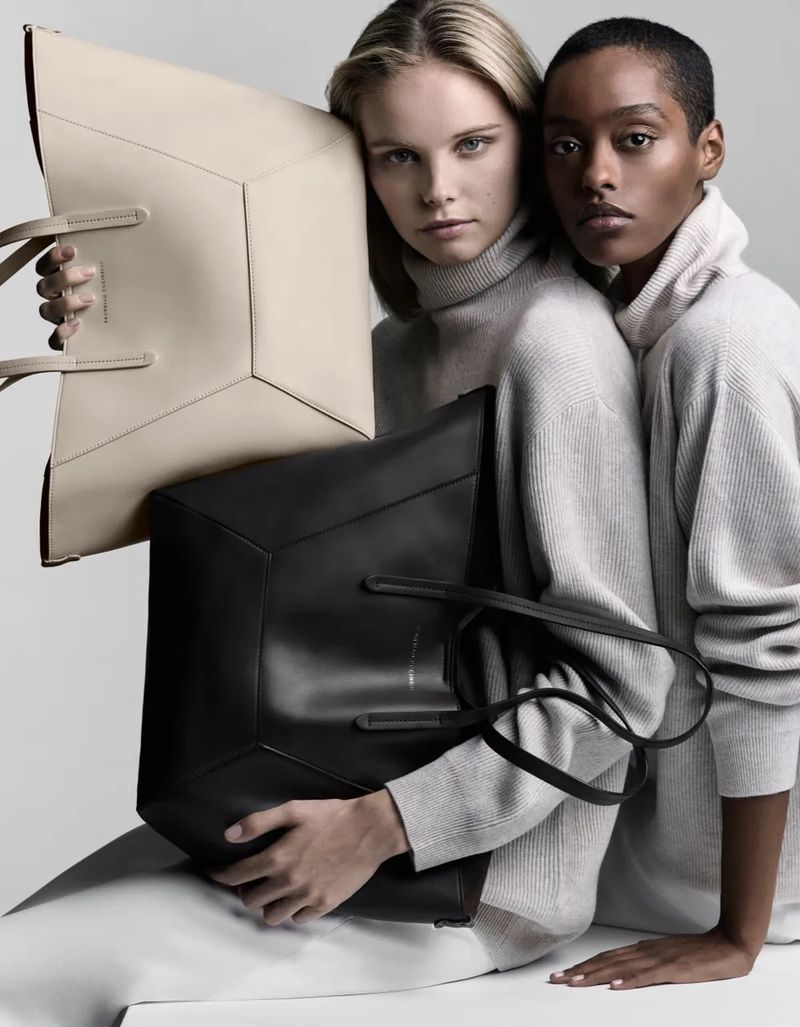 Two women pose in a studio with large geometric tote bags in black and beige. They wear light gray turtlenecks, minimal makeup.